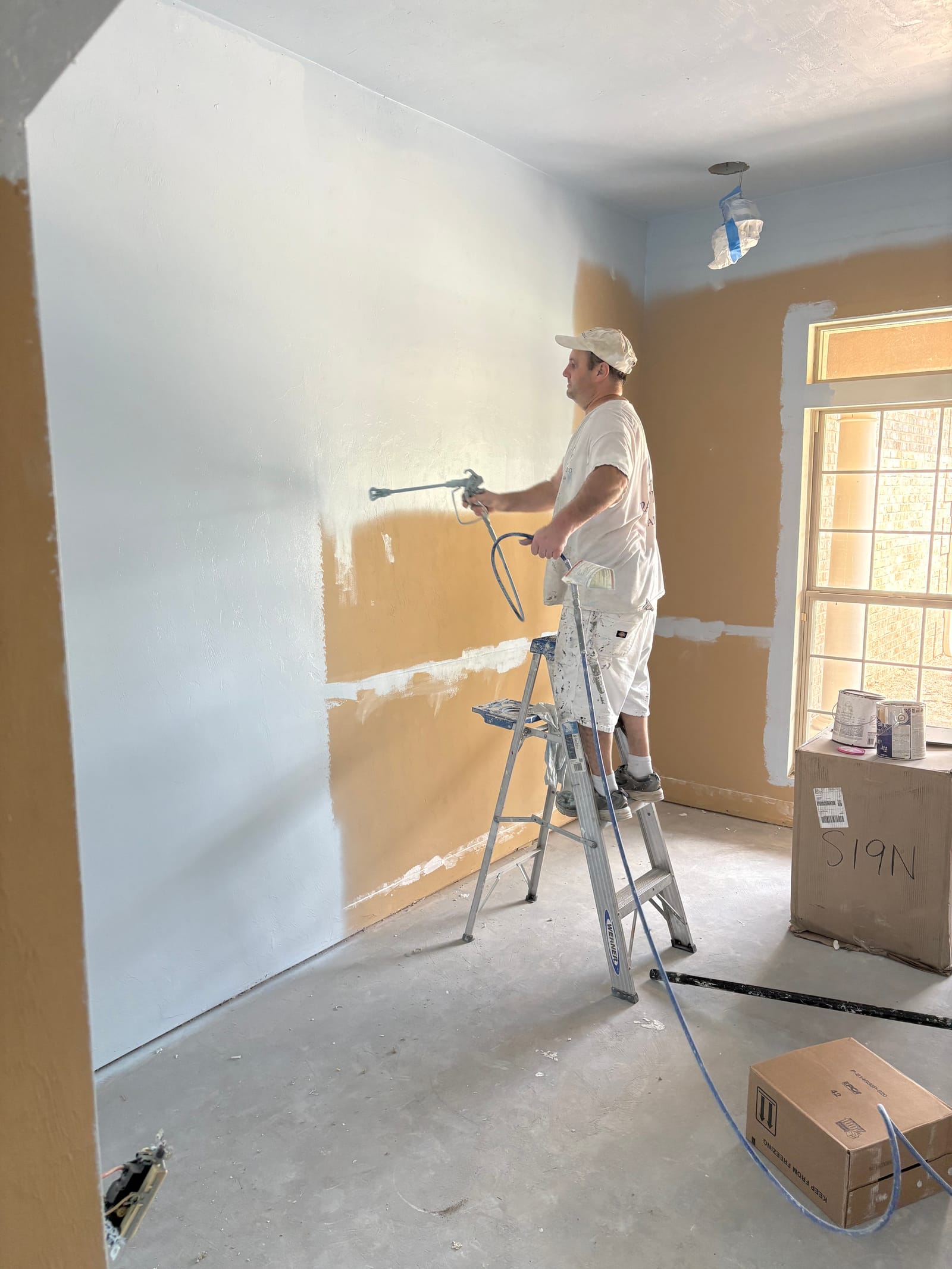 Chris with an airless paint sprayer mid-pass on an interior wall, window light behind him